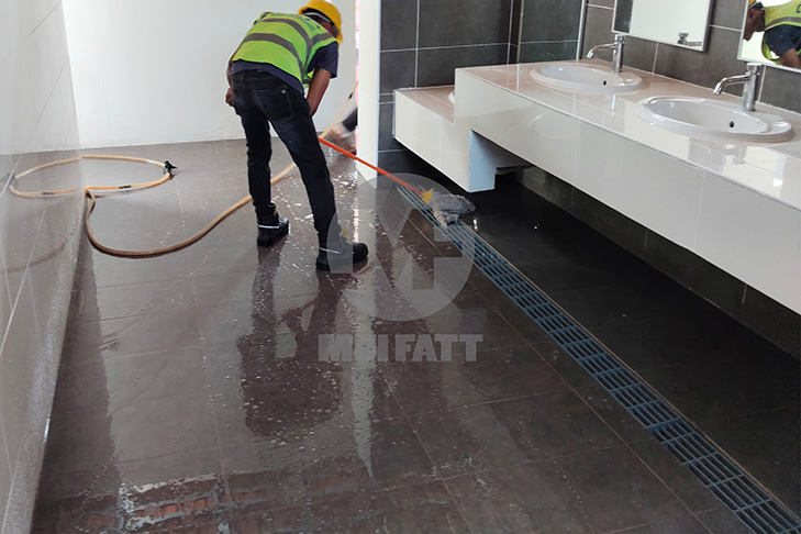 Worker washing tiled restroom floor with grey FRP molded grating drain system at RNR Gurun after installation.